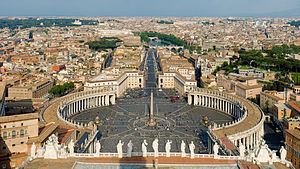 At the front of the view are the backs of thirteen large statues that stand in along the edge of the facade. Beyond them can be seen the piazza which is in three parts. The nearest appears square, while the second widens into an oval surrounded on each side by the huge grey columns on the colonnade, and with the obelisk at its centre. Beyond that is a further square surrounded by pale pink buildings. A wide street leads from the square, at the end of which can be seen the river, a bridge and castle.