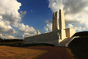 A white limestone memorial is built into the side of a hill and surrounded by green grass and backed by a blue sky. The memorial has a large front wall with rising steps on each end. Two large pylons of stone rise from a platform at the top of the wall. Human statues are located at the base of the wall on both ends, the top centre of the wall, at the base between the stone towers and near the top of the stone towers themselves.