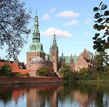A palace, with several copper-covered spires, a dome, and tiered wings with dormers, is reflected in a large artificial lake in the foreground.