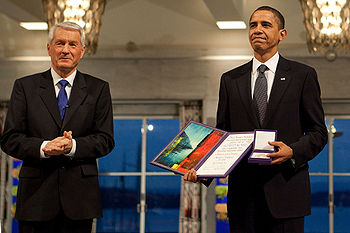 Barack Obama stands to the left of another man, both in dark suits, Obama holding his award.